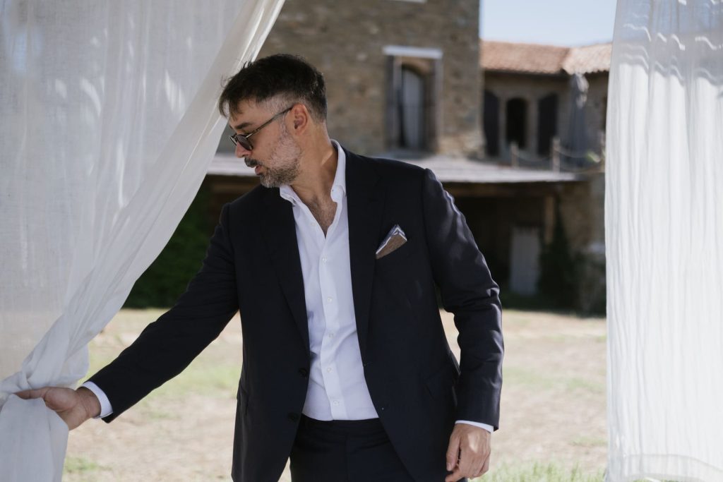 Uomo in abito scuro e occhiali da sole scosta una tenda bianca all'aperto davanti a un casale in pietra