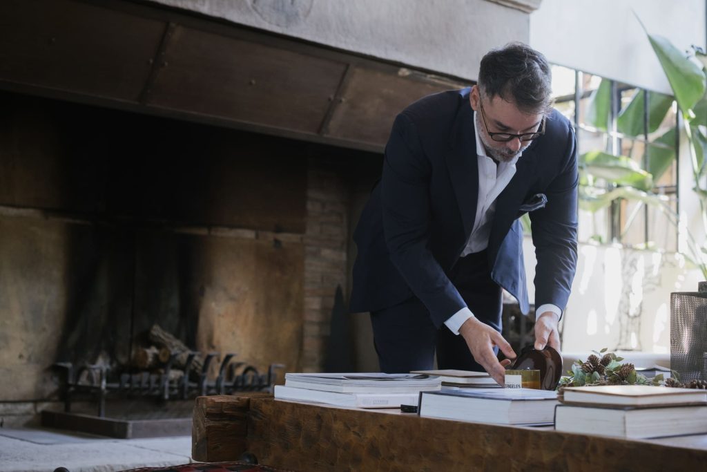 Uomo in abito scuro e occhiali si china a sistemare libri su un tavolo rustico davanti a un camino spento.